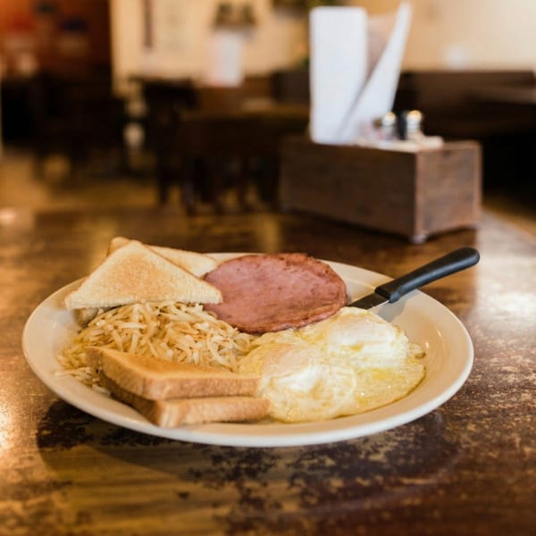 A breakfast plate with fried eggs, hash browns, ham, and toast on a rustic table.