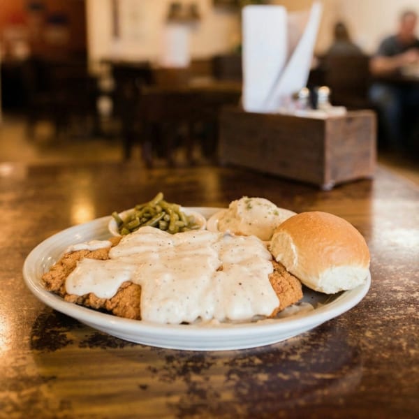 Chicken fried steak with gravy, mashed potatoes, green beans, and a dinner roll on a white plate.