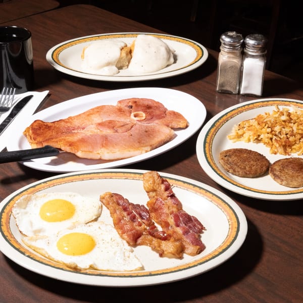 A table laden with classic diner breakfast items: biscuits and gravy, ham, hash browns, sausage, bacon, and fried eggs.
