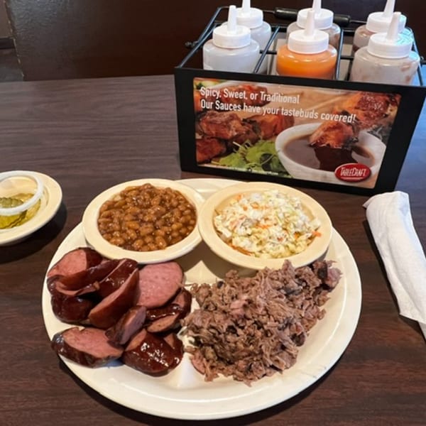 A plate of sliced sausage, pulled pork, baked beans, and coleslaw with a sauce caddy.