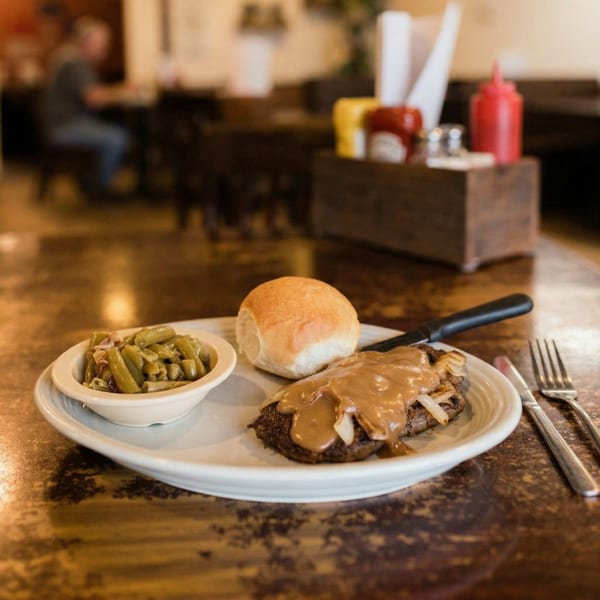 A plate of Salisbury steak with gravy and onions, green beans, and a dinner roll on a wooden table.