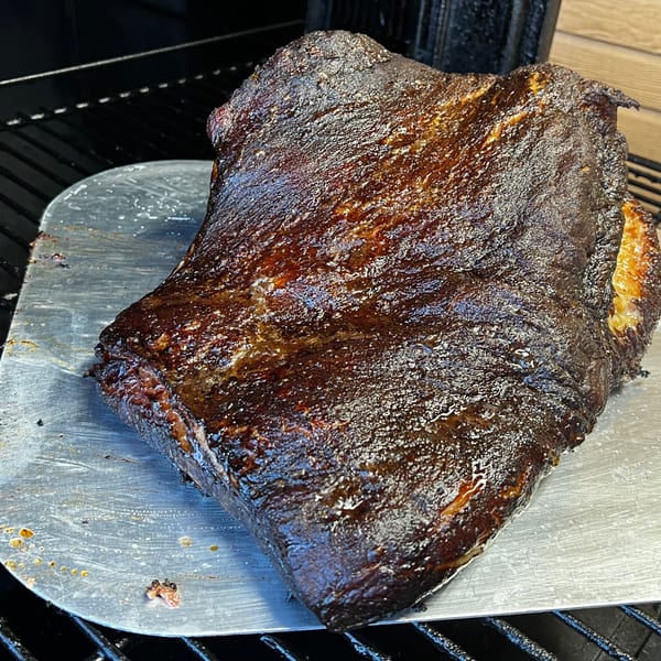 A large, dark, glistening piece of smoked meat rests on a metal peel over a grill grate.