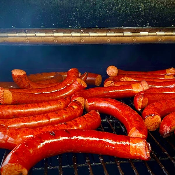 Numerous red sausages cooking on a dark grill with smoke rising from them.