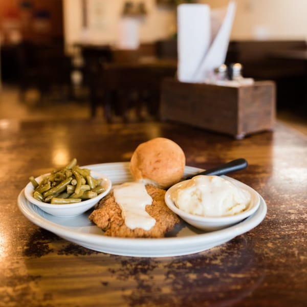 Chicken fried steak with gravy, green beans, mashed potatoes, and a roll on a wooden table.