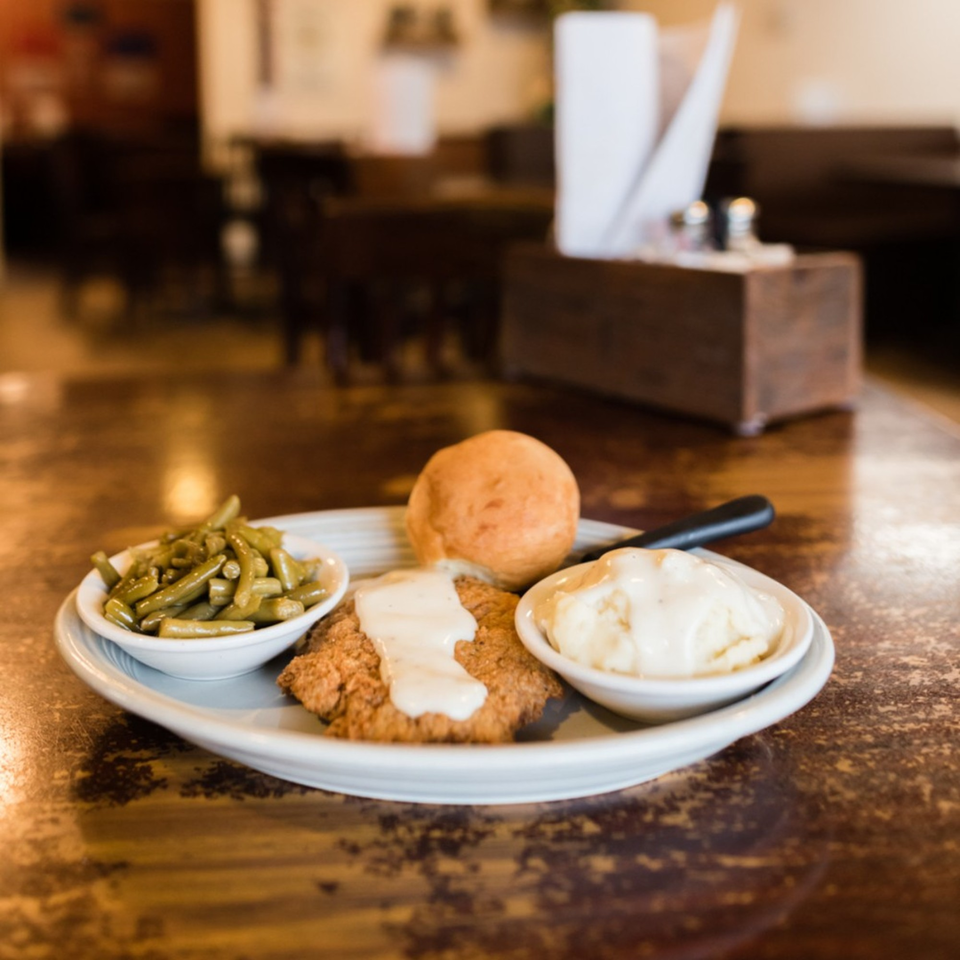 Chicken fried steak with gravy, green beans, mashed potatoes, and a roll on a wooden table.